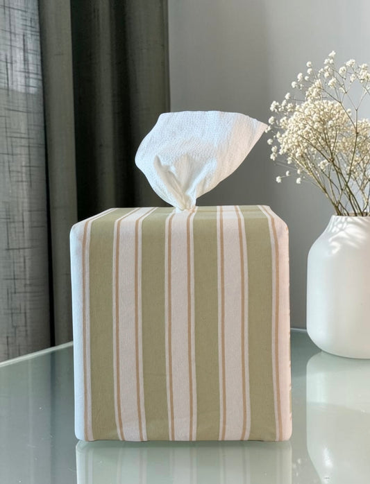 Arbor Stripe AchooBox rolled tissue dispenser vertical stripes in sage green, cream, and tan, conceals the roll of tissue inside with tissue dispensing from the top. It sits on a reflective glass table next to a white vase with dried baby's breath flowers.