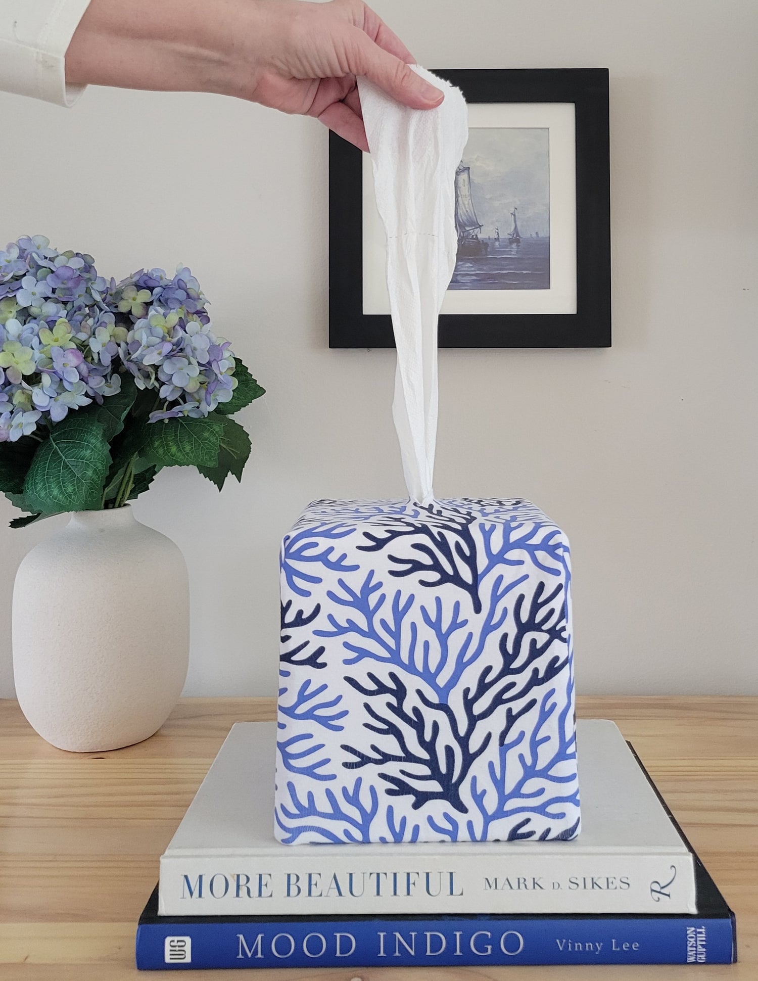Hand pulling a tissue from a Cape Cod Coral AchooBox Rolled Tissue Dispenser on books with a vase of flowers and framed picture in the background.