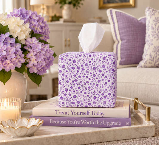 A purple floral AchooBox called Amethyst Bloom sits on stacked books on a tray in a styled living room setting with purple and white flowers, pillows, and a sofa in the background.