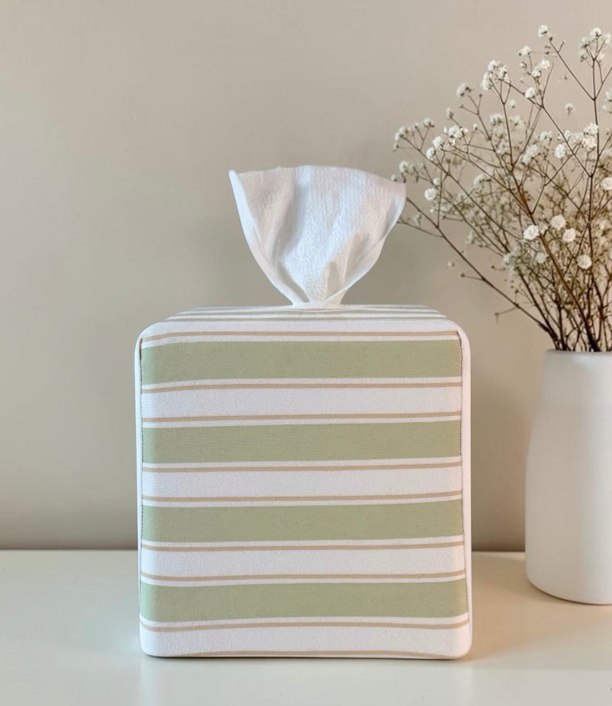 Arbor Stripe AchooBox rolled tissue dispenser has horizontal sage green and white stripes. It sits on a white surface next to a white vase with dried baby's breath flowers against a neutral beige wall.