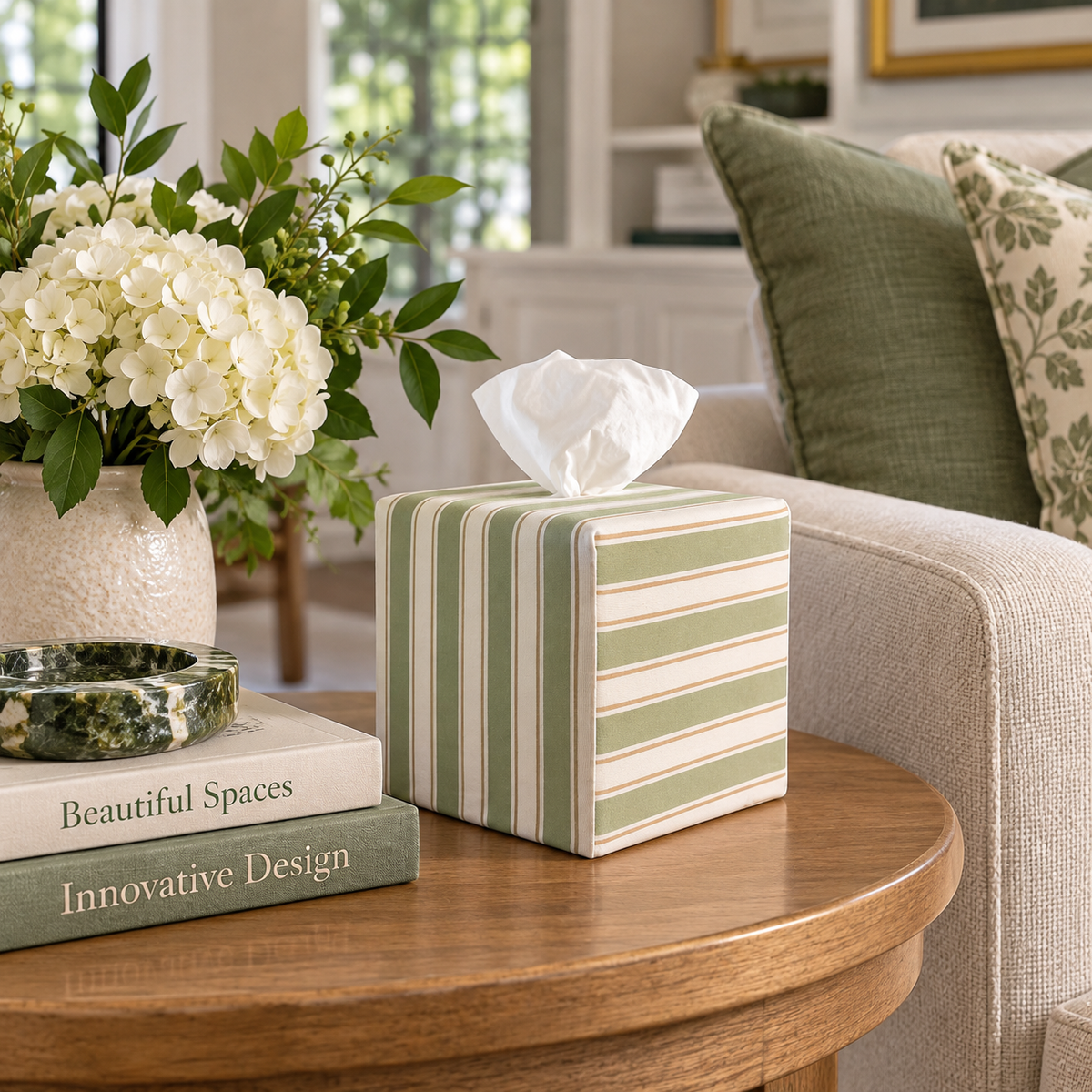 Arbor Stripe AchooBox in sage green, cream, and light brown sits on a round wooden table in a cozy living room setting next to a vase of white flowers and books.