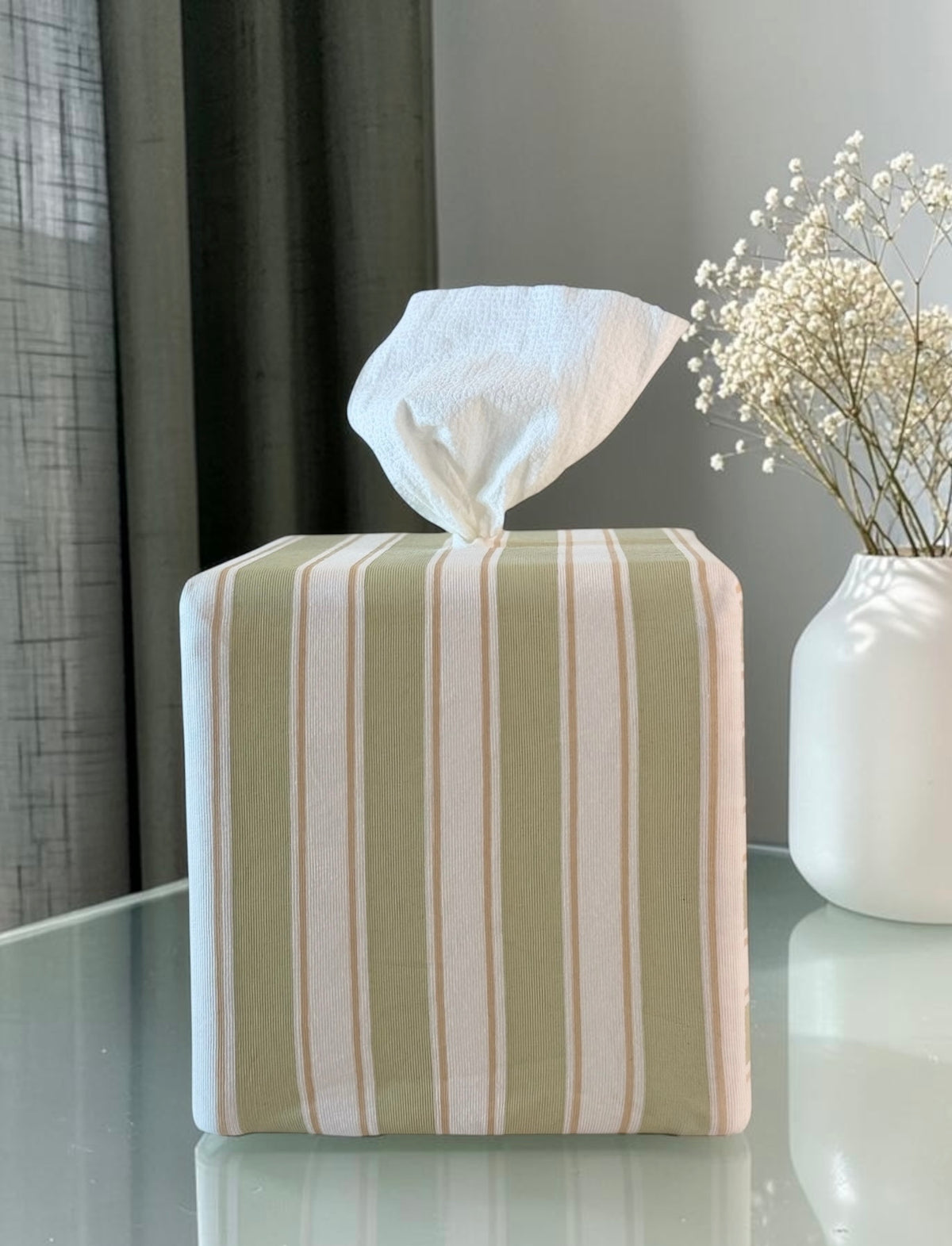 Arbor Stripe AchooBox rolled tissue dispenser vertical stripes in sage green, cream, and tan, conceals the roll of tissue inside with tissue dispensing from the top. It sits on a reflective glass table next to a white vase with dried baby's breath flowers.