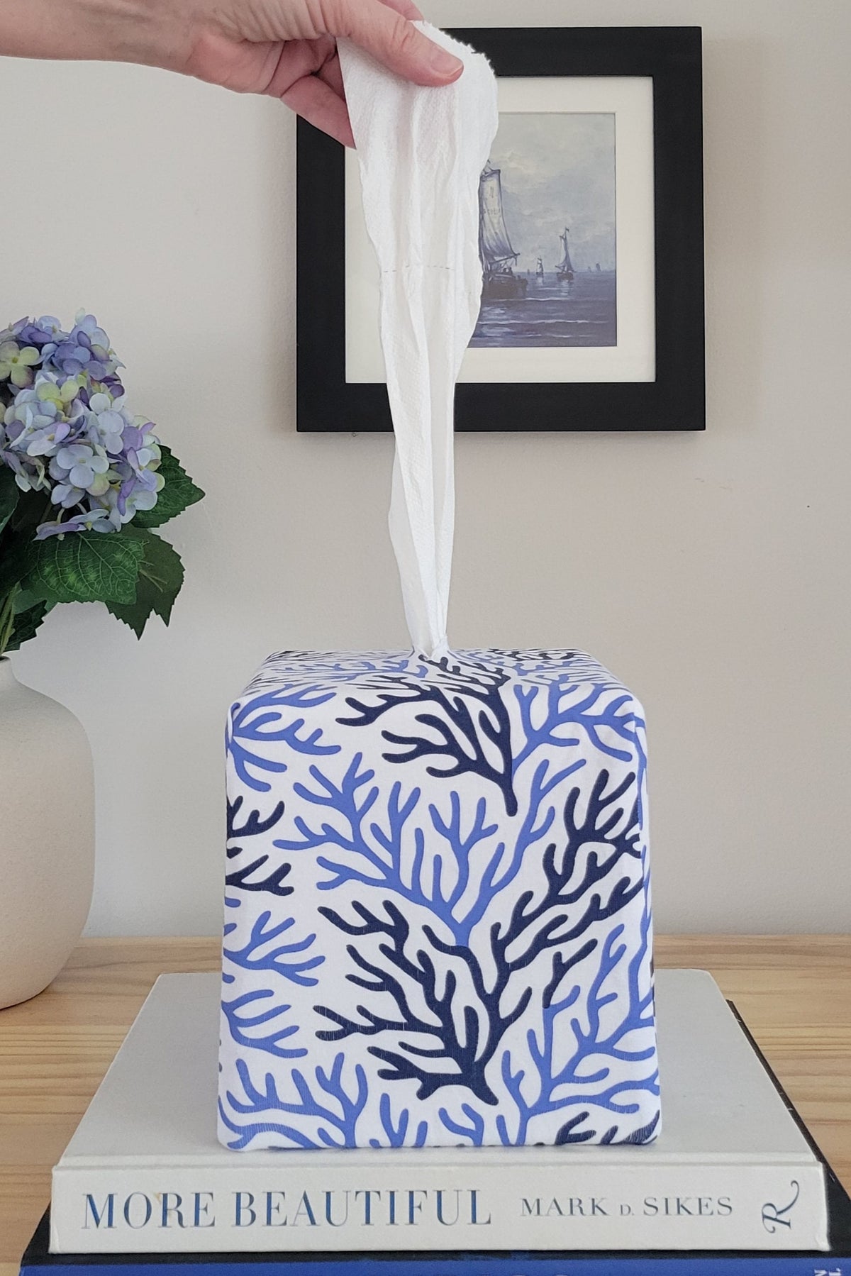 Hand pulling a tissue from a Cape Cod Coral AchooBox Rolled Tissue Dispenser on books with a vase of flowers and framed picture in the background.