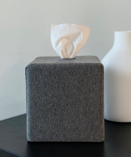 AchooBox in Charcoal solid print conceals the roll of toilet paper inside. Tissue comes out the top. It's set on a dark tabletop with a white vase blurred in the background.