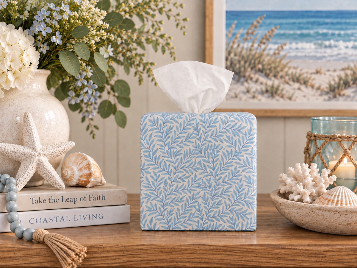 Soft Sky Leaf AchooBox rolled tissue dispenser has a light blue leaf pattern. It sits on a wooden table in a styled coastal-themed setting, surrounded by seashells, a starfish, books, and a beach scene framed on the wall.