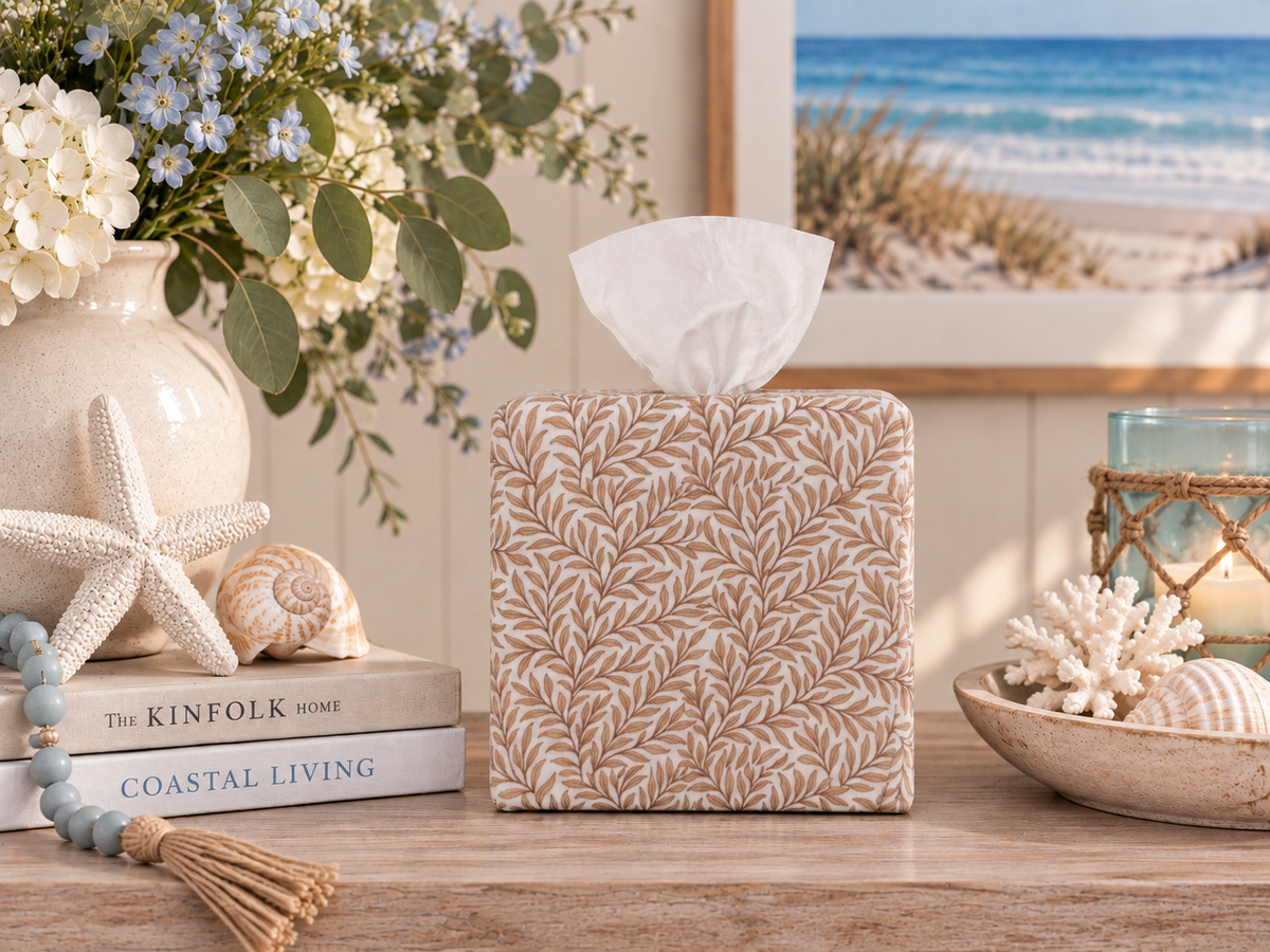 Natural Linen Leaf AchooBox has a beige and white leaf pattern. Here it sits on a wooden table in a styled coastal-themed setting, surrounded by seashells, a vase of flowers, and a framed beach print.