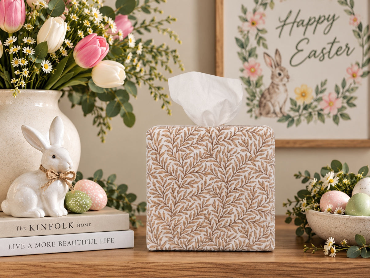 A Natural Linen Leaf AchooBox has a beige and brown leaf pattern. It sits on a wooden table in a styled Easter scene. A white rabbit figurine, a ceramic vase with tulips, pastel eggs, and an 'Happy Easter' framed print are arranged around it.