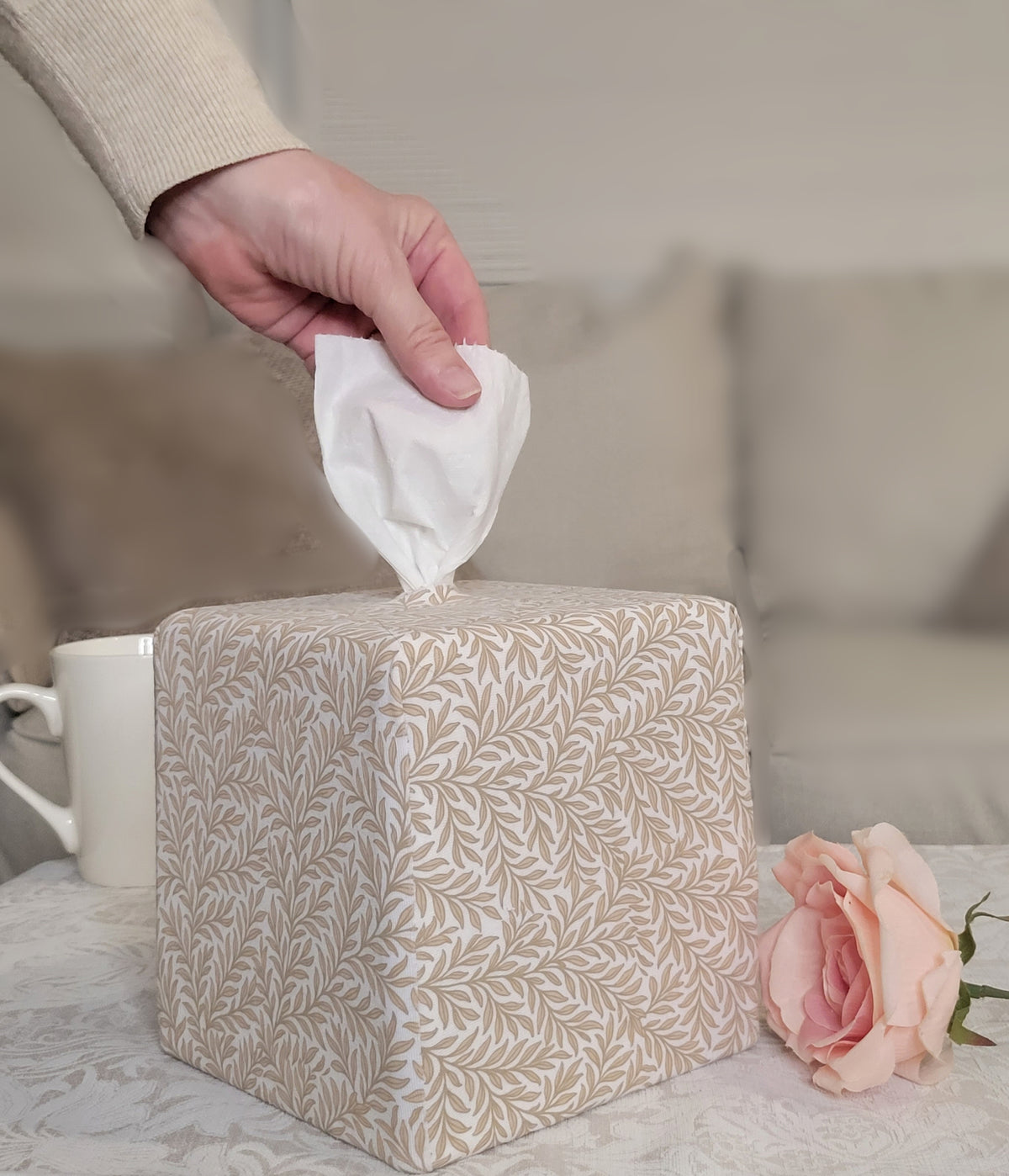 A Natural Linen Leaf AchooBox with a beige and white leaf pattern sits on a table, with a hand pulling a white tissue from the top. A pink rose and a white mug are placed beside it, with a blurred sofa in the background.