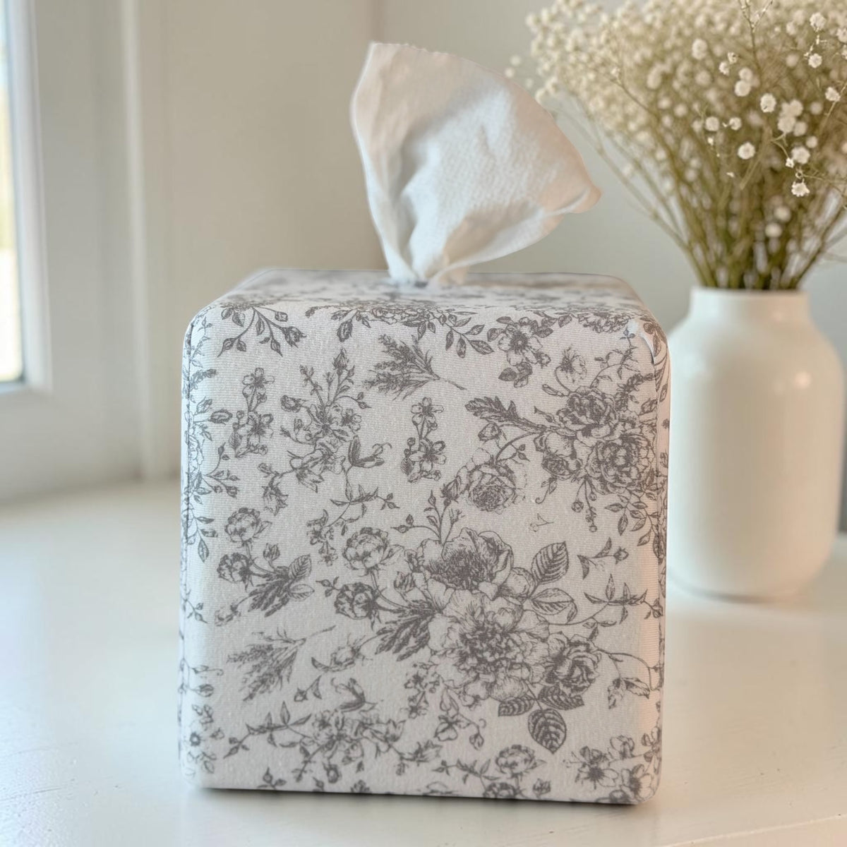 Parisian Slate Heirloom Toile AchooBox with bath tissue concealed inside and coming out of the top like a standard tissue box. White vase with baby's breath in the background.