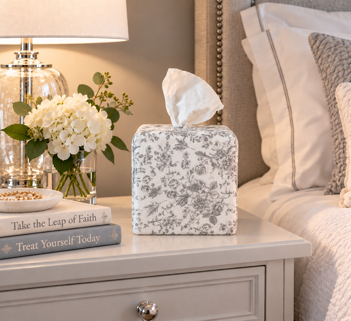 Parisian Slate Heirloom Toile AchooBox on a light gray nightstand next to a couple of books Take the Leap of Faith and Treat Yourself Today and a glass vase with white hydrangea