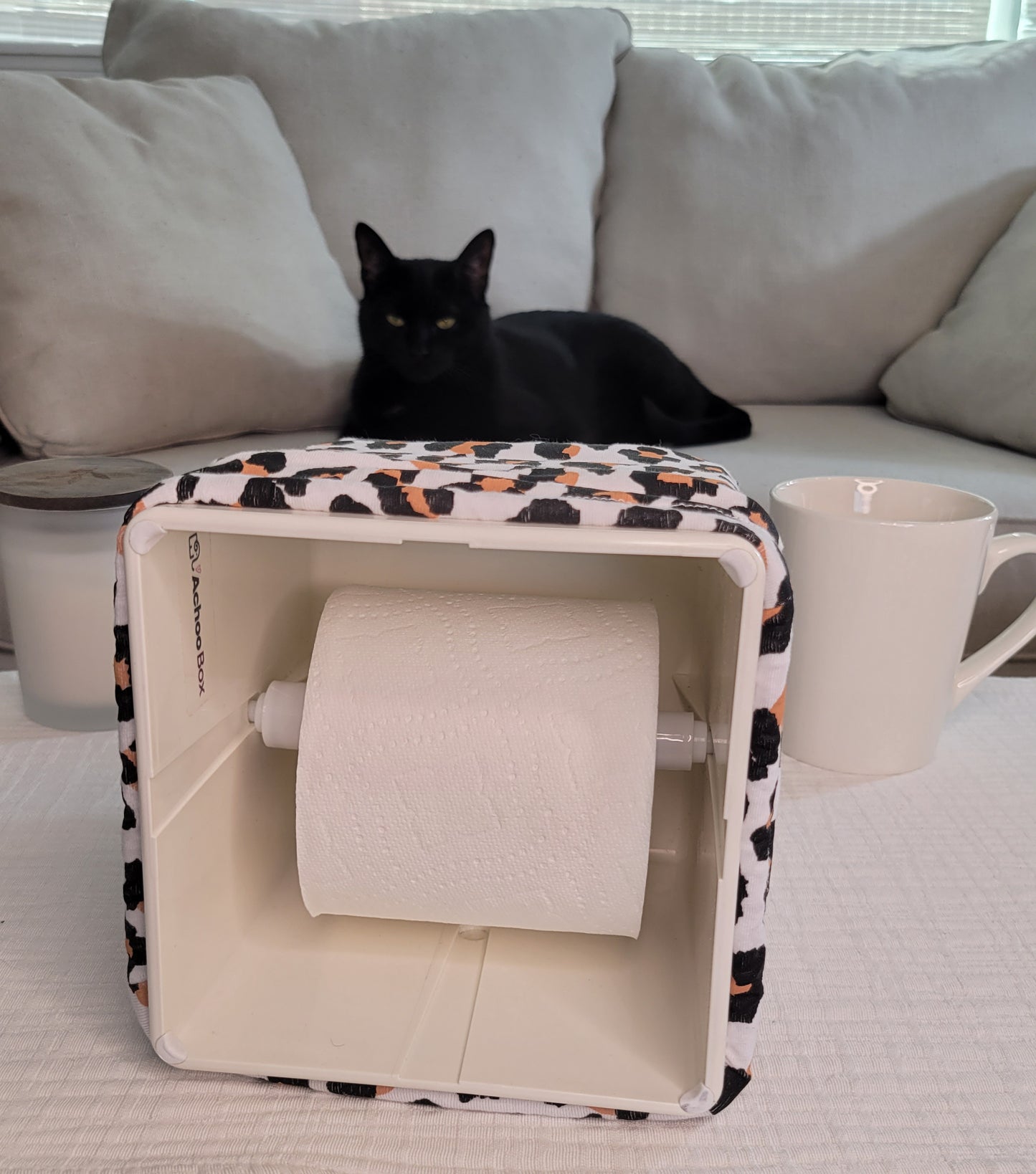 Modern Leopard AchooBox tipped on its side to show the roll of tissue with Merlin the Cat sitting on the couch.