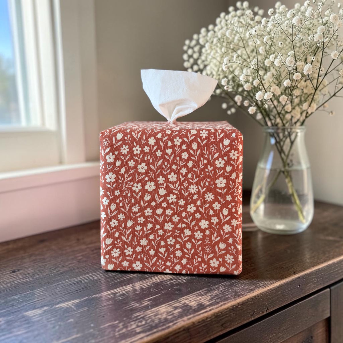 Rooibos Tea Bloom AchooBox with a white floral pattern on a terracotta red background, sitting on a dark wooden table next to a vase of baby's breath. 