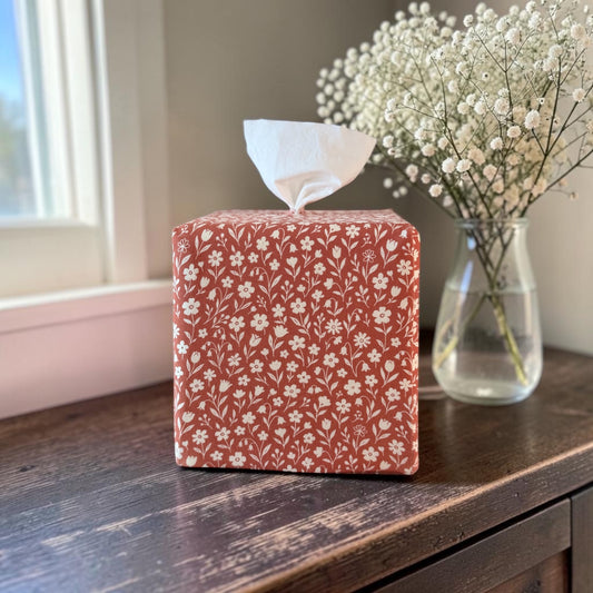 Rooibos Tea Bloom AchooBox with a white floral pattern on a terracotta red background, sitting on a dark wooden table next to a vase of baby's breath. 