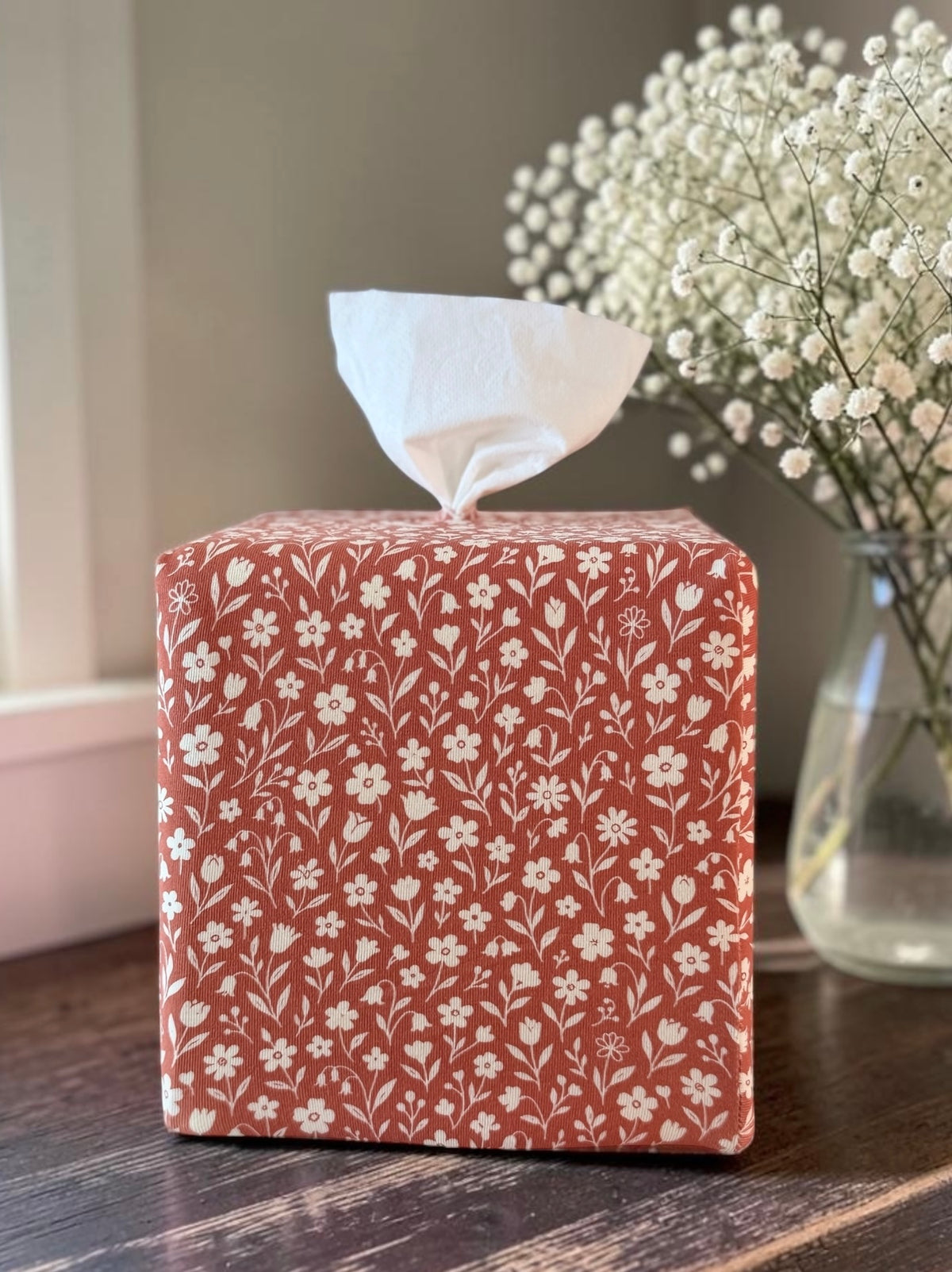 AchooBox with Rooibos Tea AchooCover is a terracotta red and white floral pattern. It sits on a dark wooden table, with a white tissue pulled out of the top and baby's breath flowers in a glass vase blurred in the background.