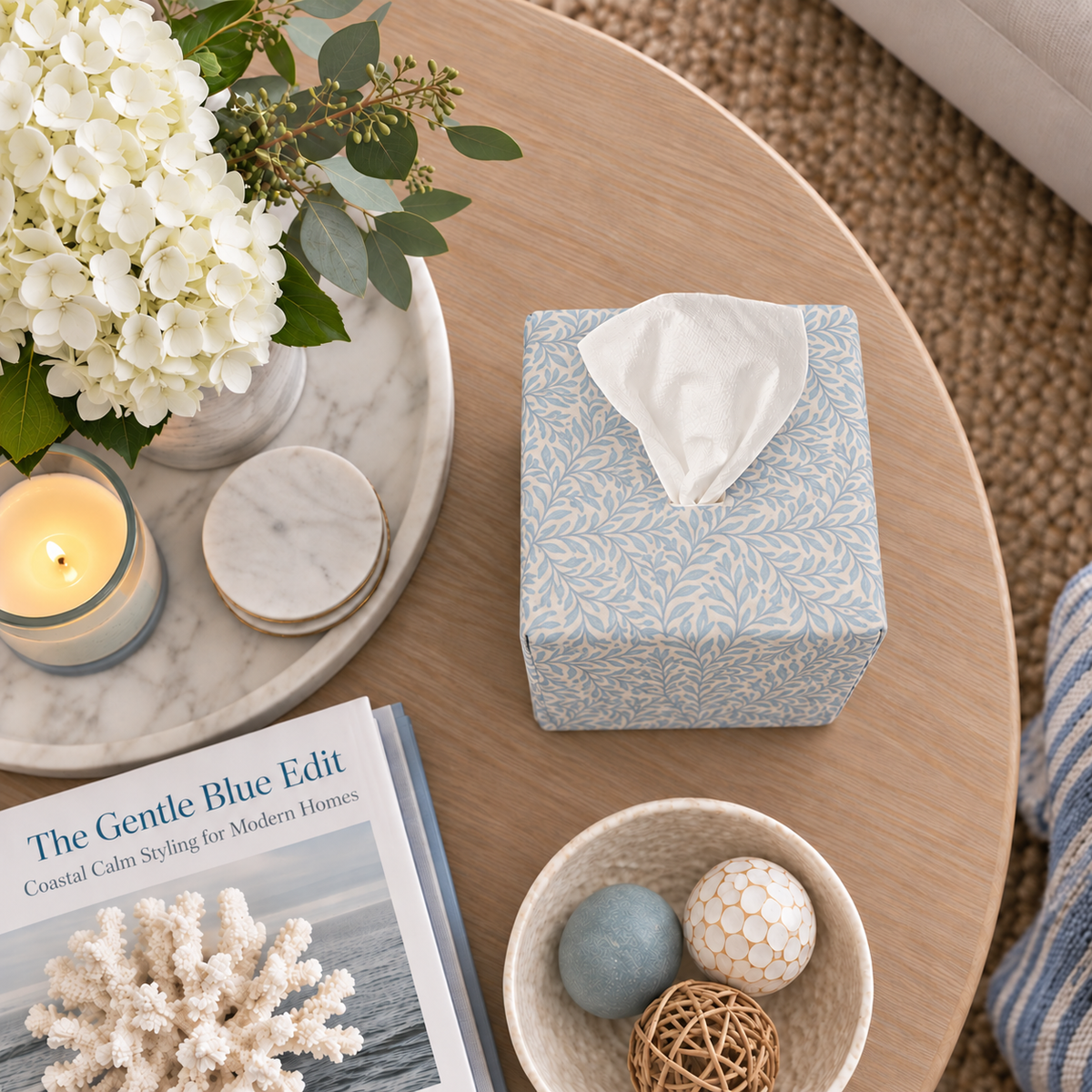 Soft Sky Leaf AchooBox with Tissue coming out of the top. On a round coffee table with some coastal themed elements such as coral sitting on a book The Gentle Blue Edit