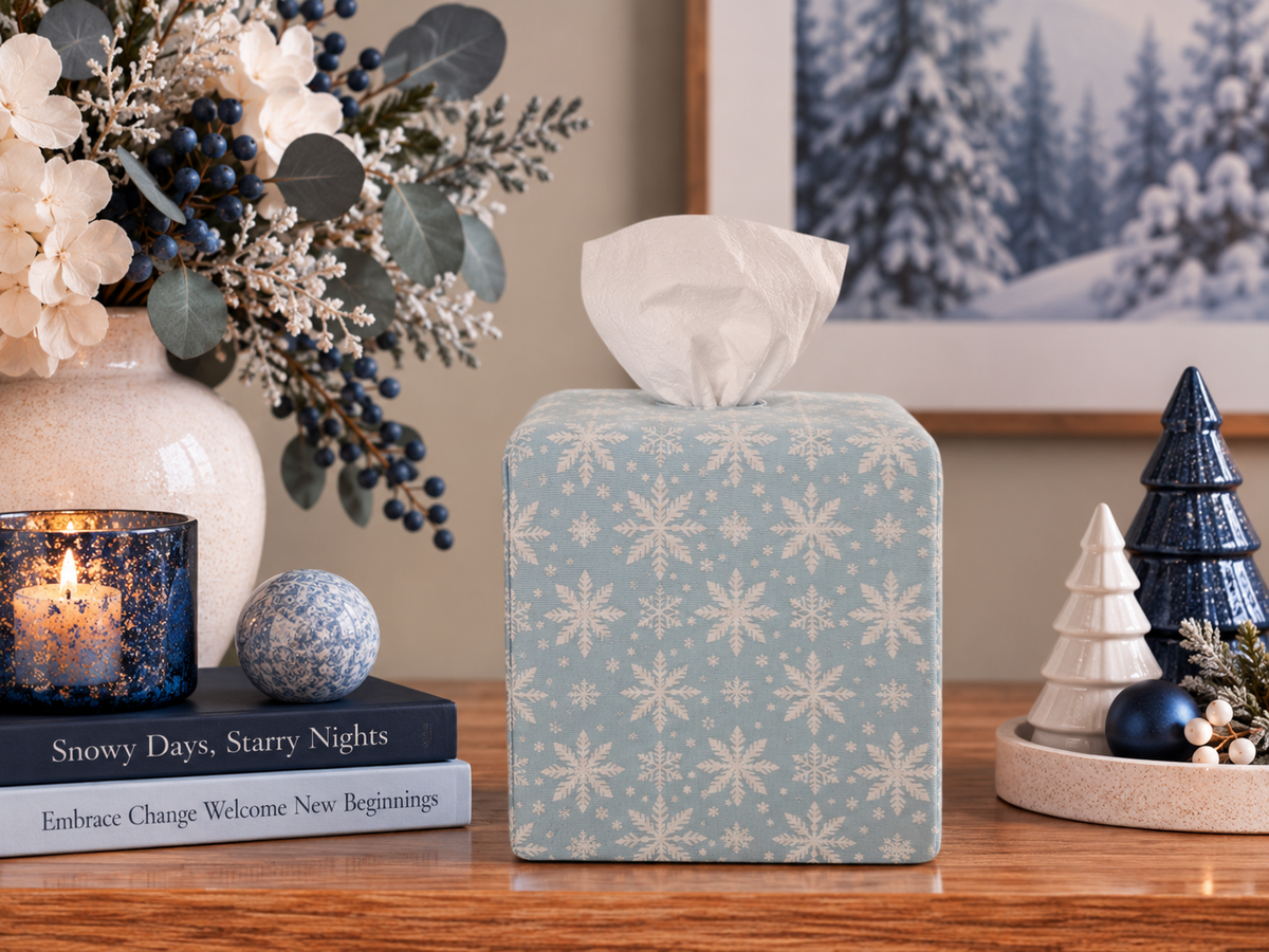 AchooBox with Winter Snow Achoocover - a light blue fabric with a white snowflake pattern, placed on a wooden table in a styled holiday setting with a candle, books, and decorative trees.
