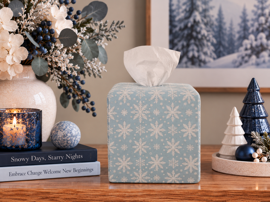 AchooBox with Winter Snow Achoocover - a light blue fabric with a white snowflake pattern, placed on a wooden table in a styled holiday setting with a candle, books, and decorative trees.